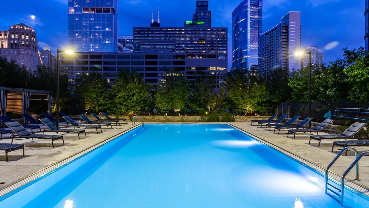 Illuminated outdoor swimming pool at dusk, surrounded by lounge chairs with a vibrant Chicago city skyline at Hubbard Place