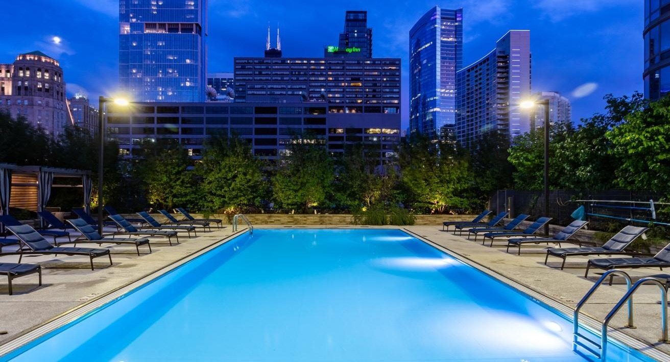 Illuminated outdoor swimming pool at dusk, surrounded by lounge chairs with a vibrant Chicago city skyline at Hubbard Place