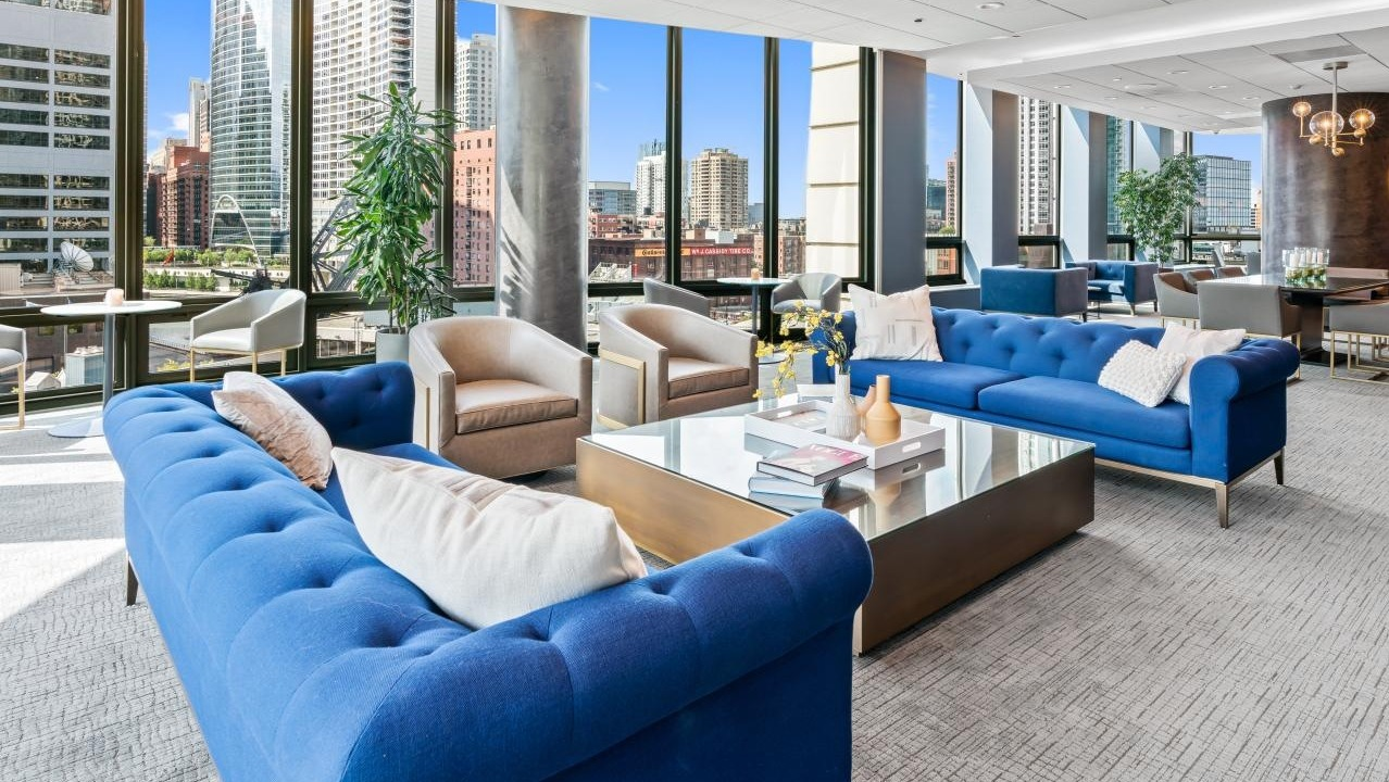 Expansive resident lounge with vibrant blue sofas, comfortable chairs, and panoramic city skyline views at Hubbard Place in Chicago