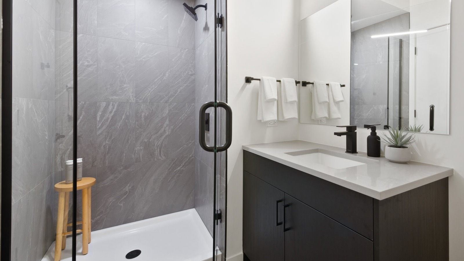 Contemporary bathroom featuring a dark vanity, modern mirror, and a spacious walk-in shower with grey tiles at Hugo Apartments River North, Chicago