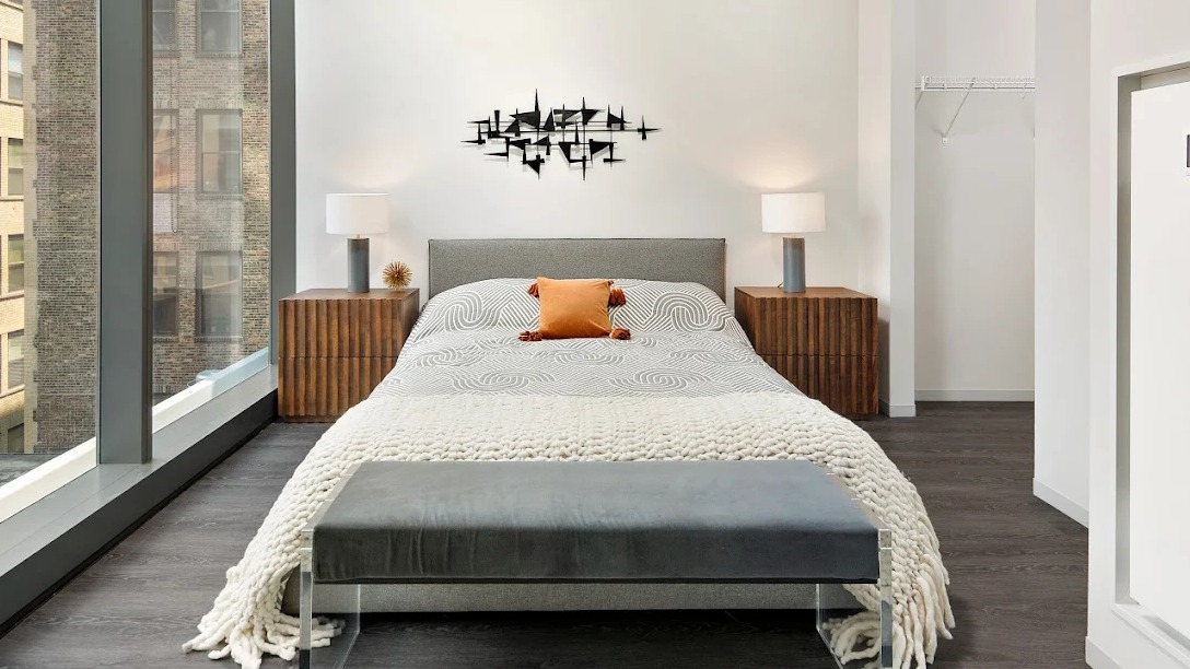 Modern bedroom with a grey bed, wooden nightstands, a stylish bench, and large windows at Imprint apartments in Chicago