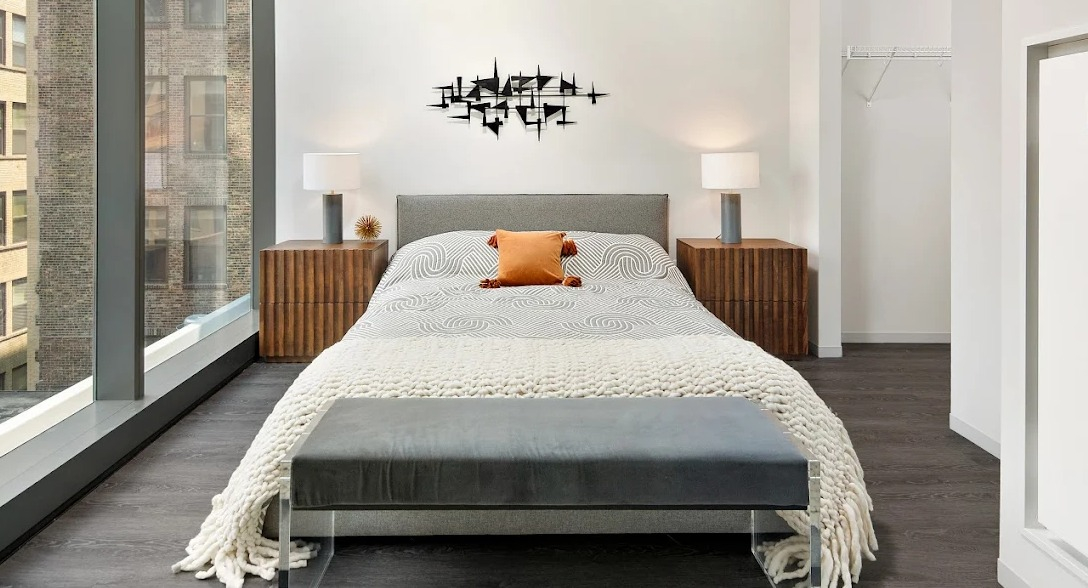 Modern bedroom with a grey bed, wooden nightstands, a stylish bench, and large windows at Imprint apartments in Chicago