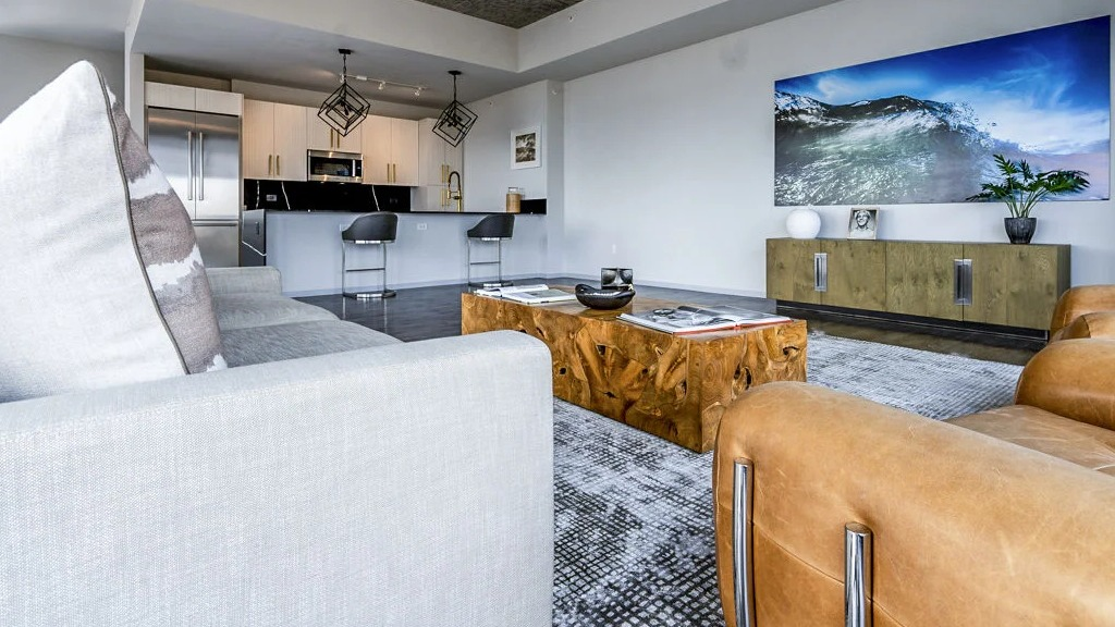Contemporary kitchen and living room with a grey sofa, leather chairs, a large TV, and an island with bar seating at Imprint apartments in Chicago