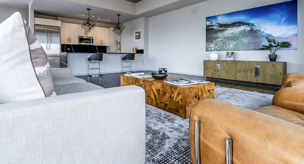 Contemporary kitchen and living room with a grey sofa, leather chairs, a large TV, and an island with bar seating at Imprint apartments in Chicago
