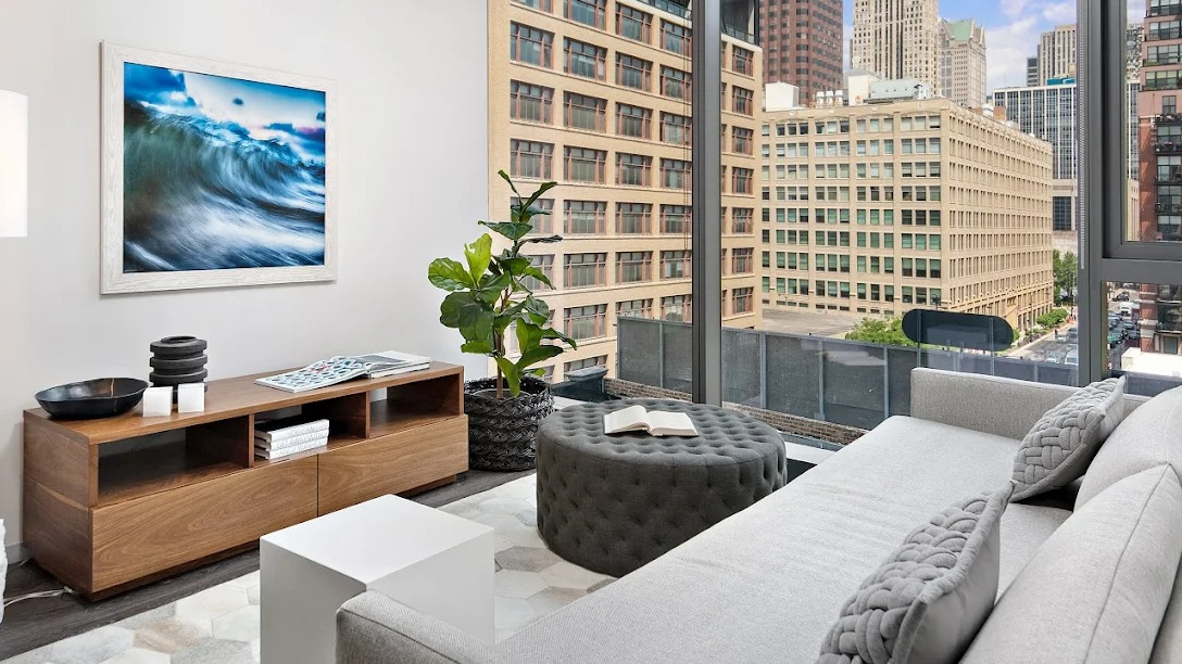Contemporary living room with a grey sofa, ottoman, wooden media console, and large windows with city views at Imprint apartments in Chicago