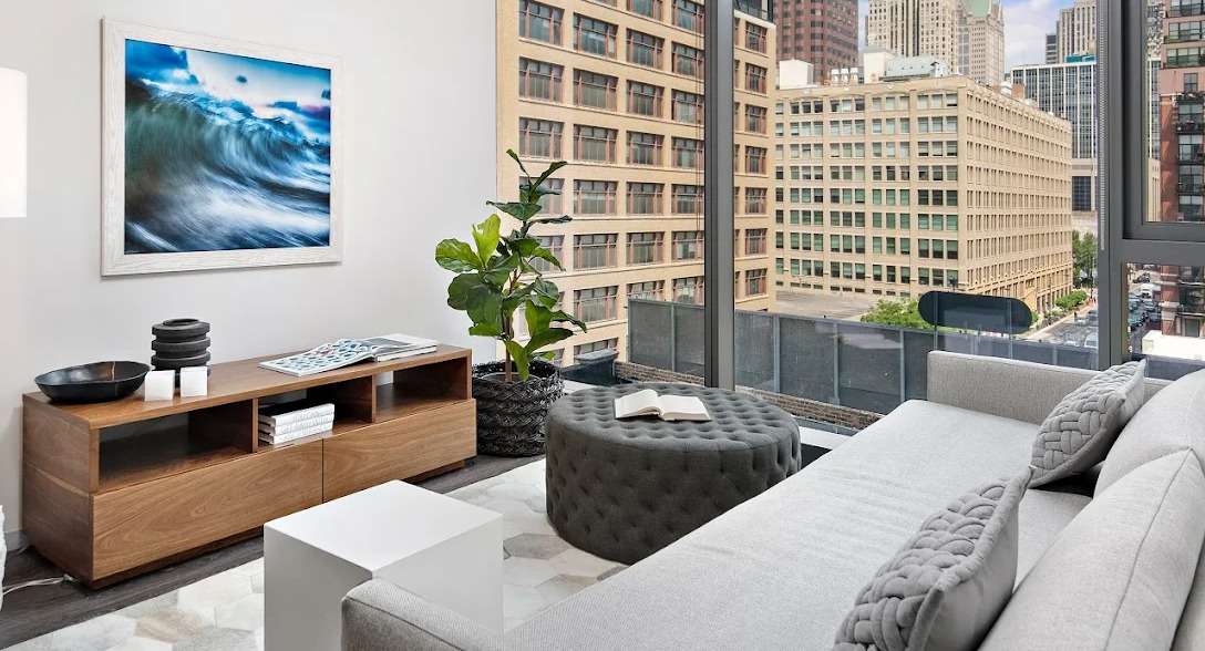 Contemporary living room with a grey sofa, ottoman, wooden media console, and large windows with city views at Imprint apartments in Chicago