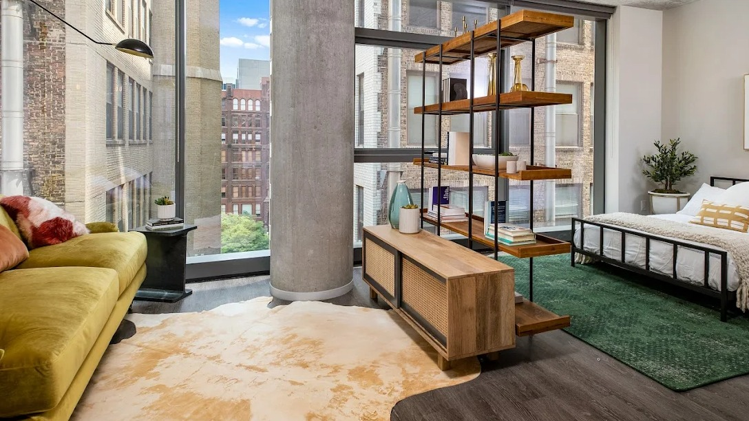 Stylish studio apartment layout featuring a green sofa, bookshelf room divider, bed, and large windows at Imprint apartments in Chicago