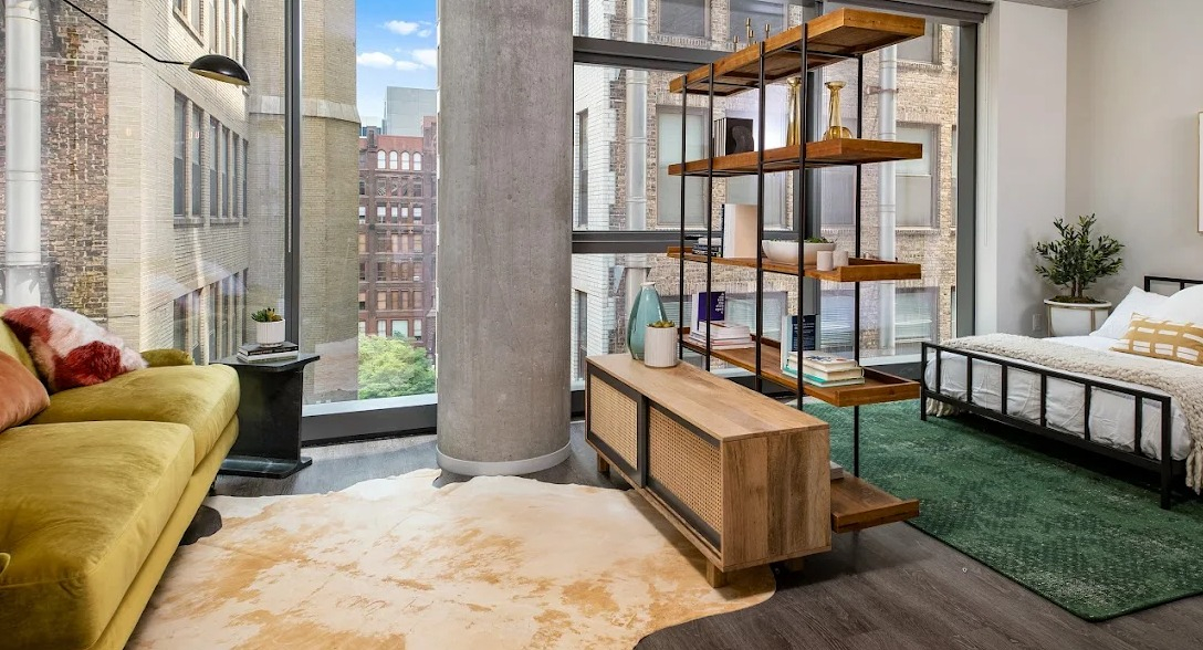 Stylish studio apartment layout featuring a green sofa, bookshelf room divider, bed, and large windows at Imprint apartments in Chicago