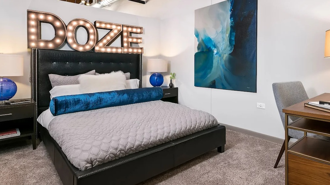Unique bedroom with a large bed, stylish "DOZE" light-up sign, blue accent decor, and a desk area at Lofts at River East in Chicago