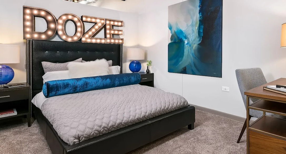 Unique bedroom with a large bed, stylish "DOZE" light-up sign, blue accent decor, and a desk area at Lofts at River East in Chicago