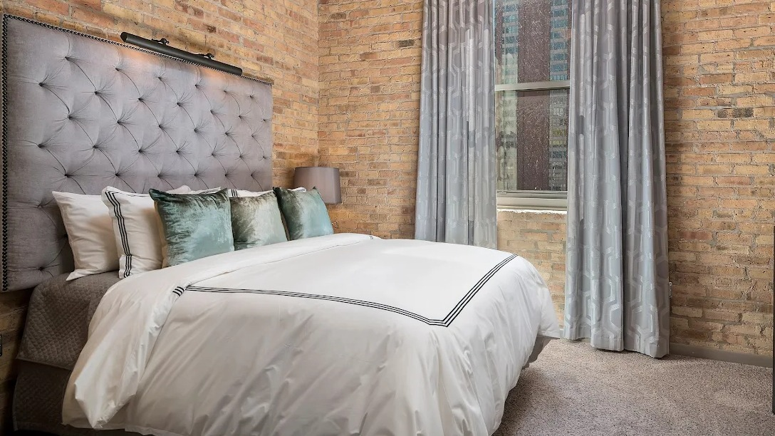 Cozy bedroom featuring exposed brick walls, a comfortable bed with a tufted headboard, and soft lighting at Lofts at River East in Chicago