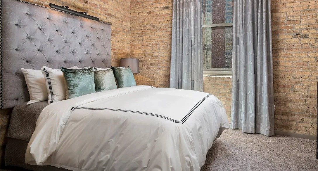 Cozy bedroom featuring exposed brick walls, a comfortable bed with a tufted headboard, and soft lighting at Lofts at River East in Chicago