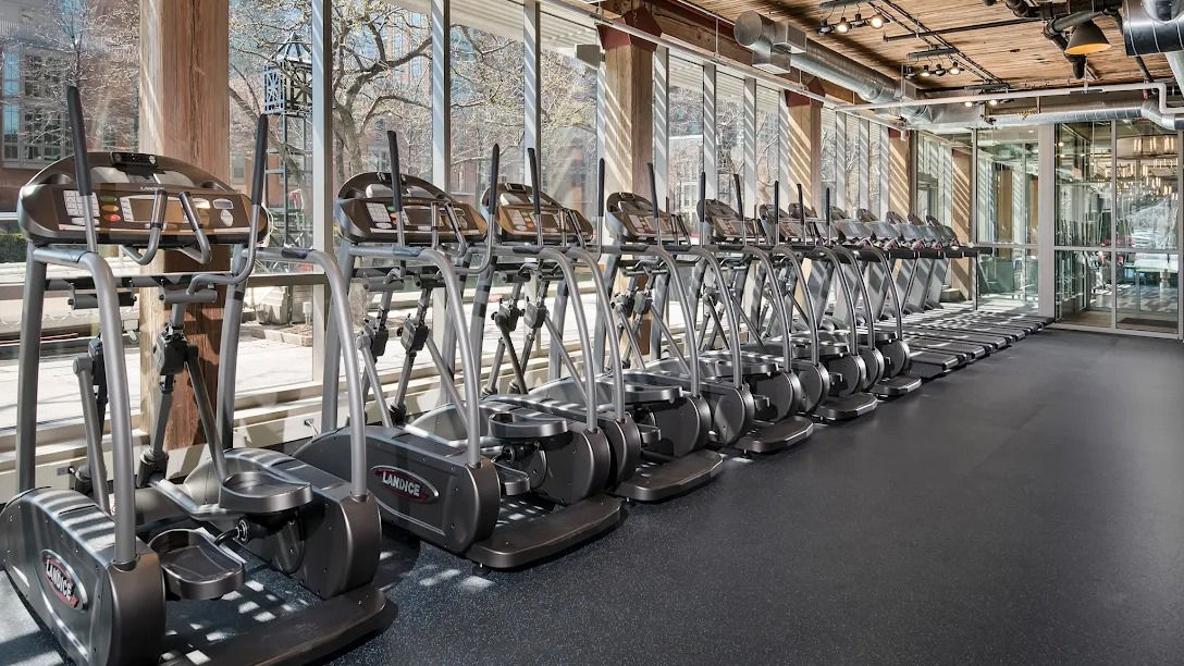 Bright cardio room with a row of ellipticals, large windows, and exposed wood and ductwork ceilings at Lofts at River East in Chicago