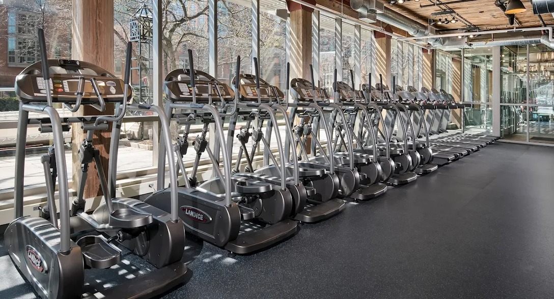 Bright cardio room with a row of ellipticals, large windows, and exposed wood and ductwork ceilings at Lofts at River East in Chicago