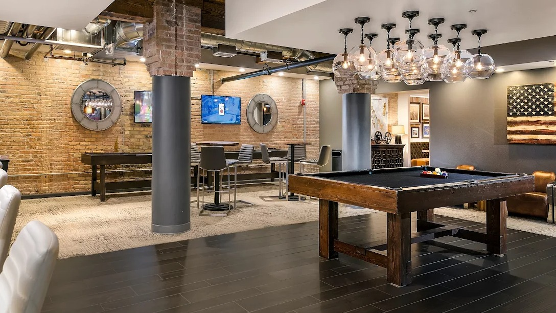Lively game room featuring a pool table, bar area with seating, arcade games, and exposed brick walls at Lofts at River East in Chicago