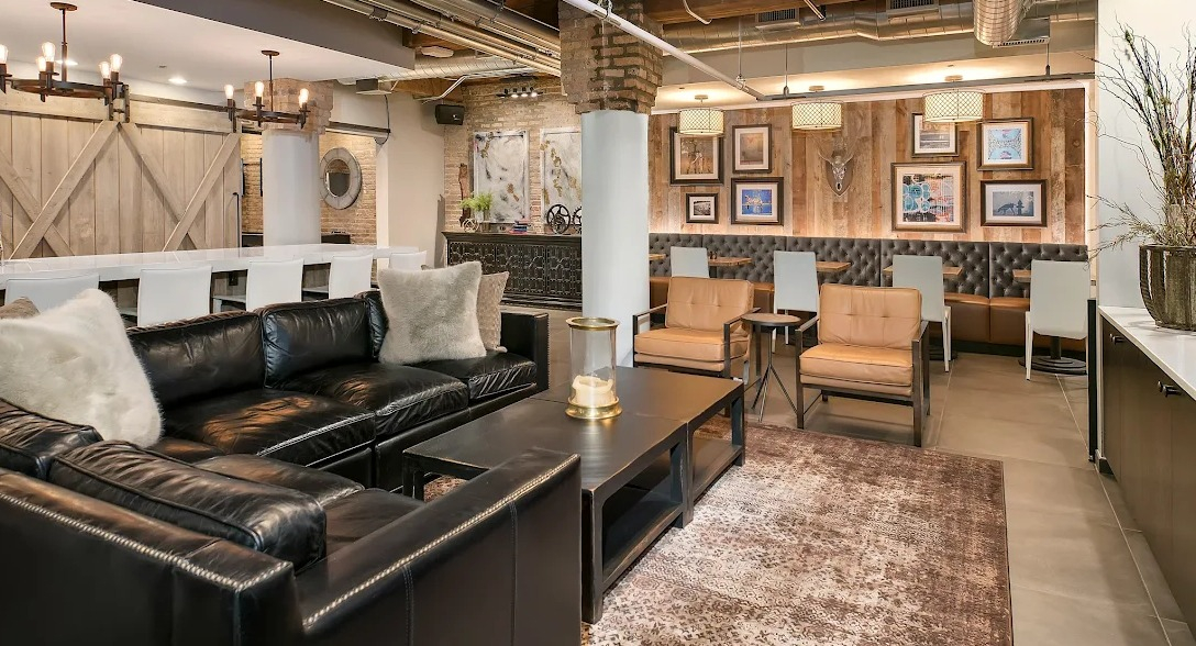Rustic-chic resident lounge with leather sofas, exposed brick, a long bar, and framed artwork, creating a cozy social space at Lofts at River East in Chicago