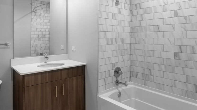 A clean bathroom at Luxe on Chicago Apartments featuring a wood vanity, white tiled shower, and a pristine bathtub