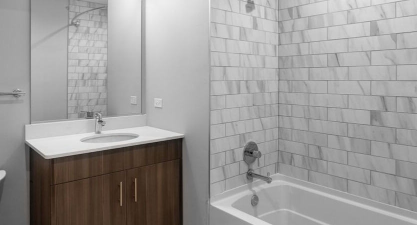 A clean bathroom at Luxe on Chicago Apartments featuring a wood vanity, white tiled shower, and a pristine bathtub