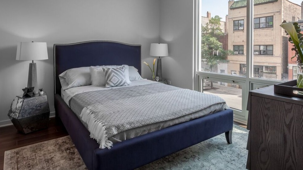 A cozy bedroom at Luxe on Chicago Apartments with a blue upholstered bed and large windows providing city views