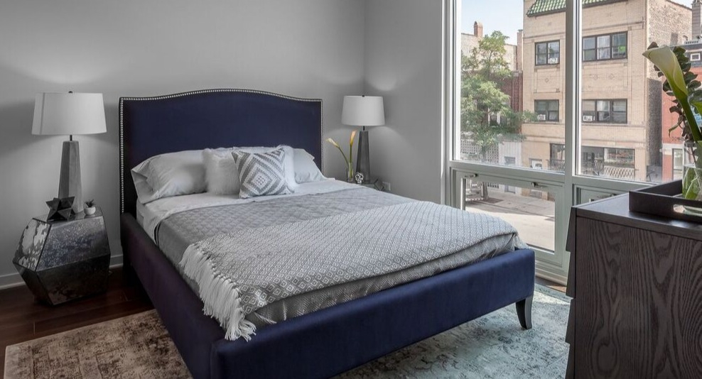 A cozy bedroom at Luxe on Chicago Apartments with a blue upholstered bed and large windows providing city views