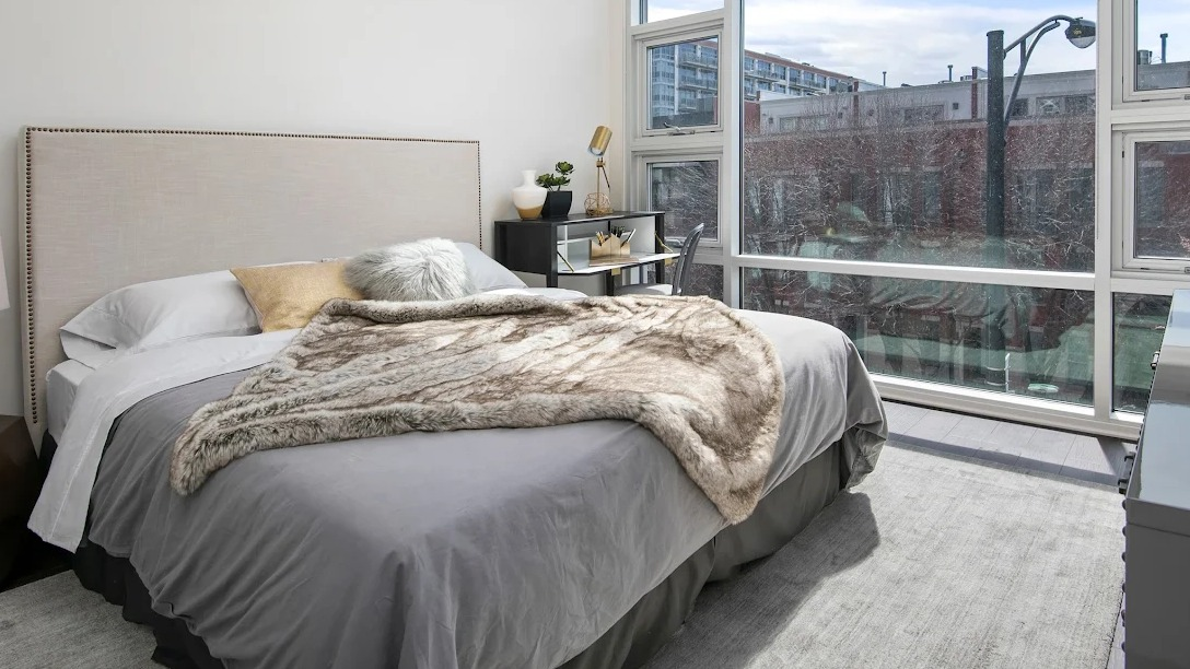 Elegant bedroom with a plush bed, modern desk, and large windows offering urban views at Luxe on Madison Apartments in Chicago