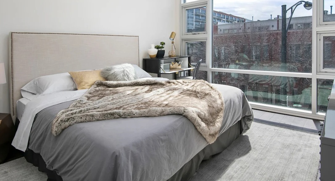 Elegant bedroom with a plush bed, modern desk, and large windows offering urban views at Luxe on Madison Apartments in Chicago