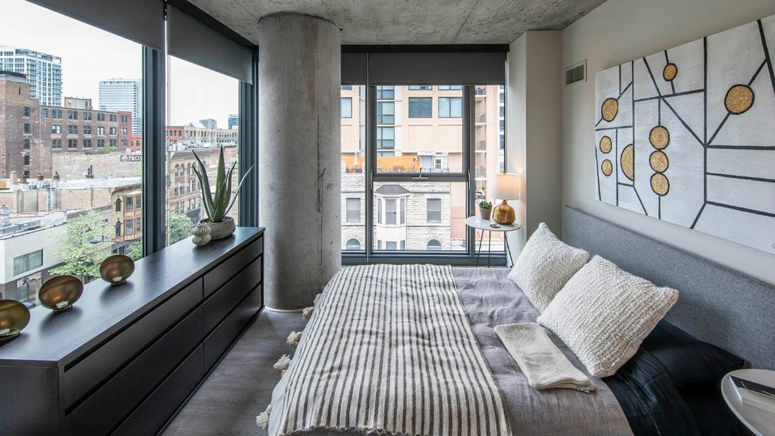 Modern bedroom with exposed concrete ceiling, a comfortable bed, stylish decor, and large windows with urban views at Marlowe Apartments in Chicago