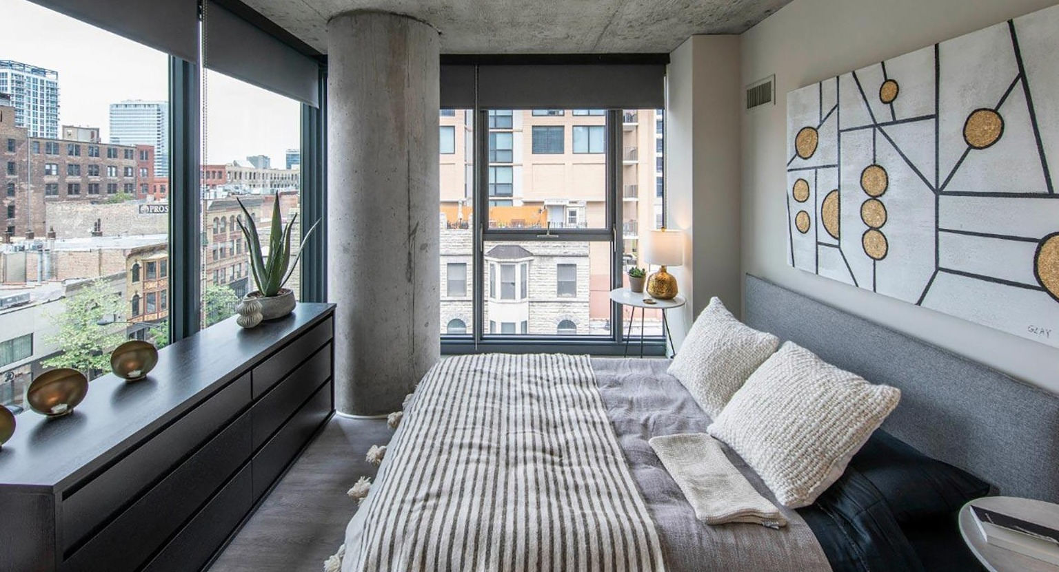 Modern bedroom with exposed concrete ceiling, a comfortable bed, stylish decor, and large windows with urban views at Marlowe Apartments in Chicago