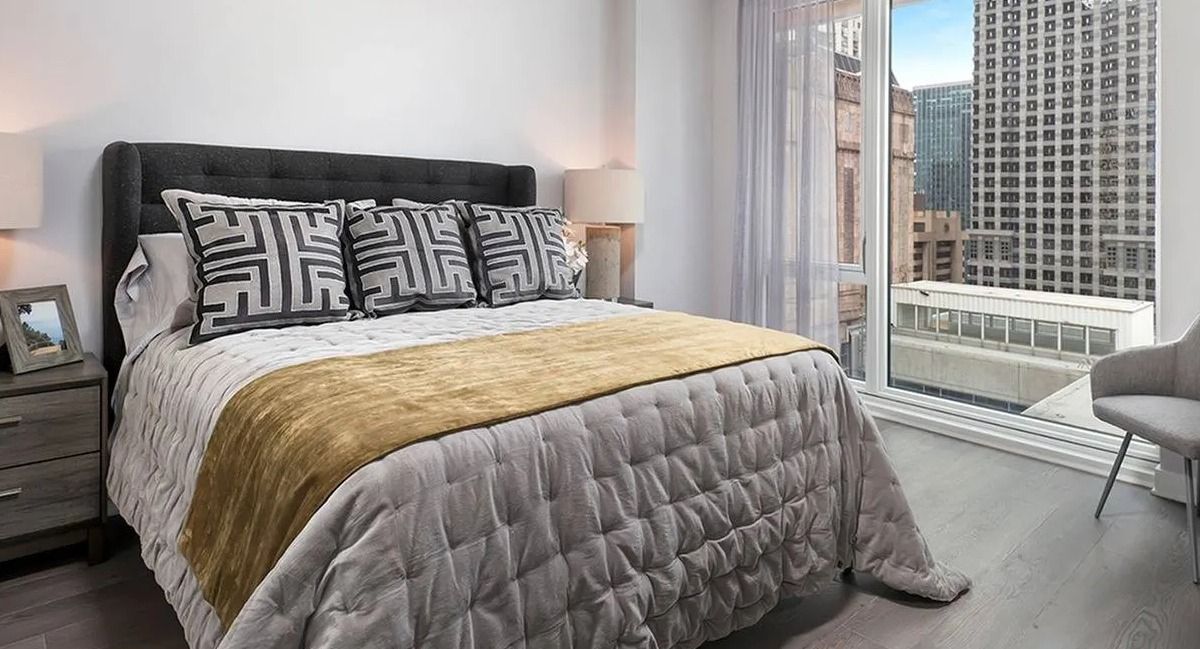 Contemporary bedroom with a large bed, elegant grey and gold bedding, and city views from expansive windows at Marquee at Block 37 in Chicago