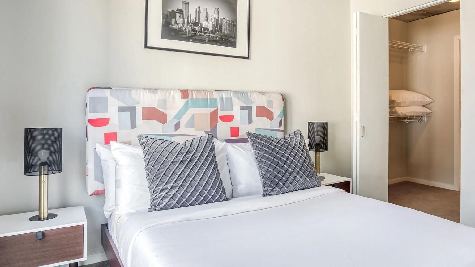 Contemporary bedroom with a patterned headboard, bedside tables, and an open closet showing shelving at MDA Apartments in Chicago