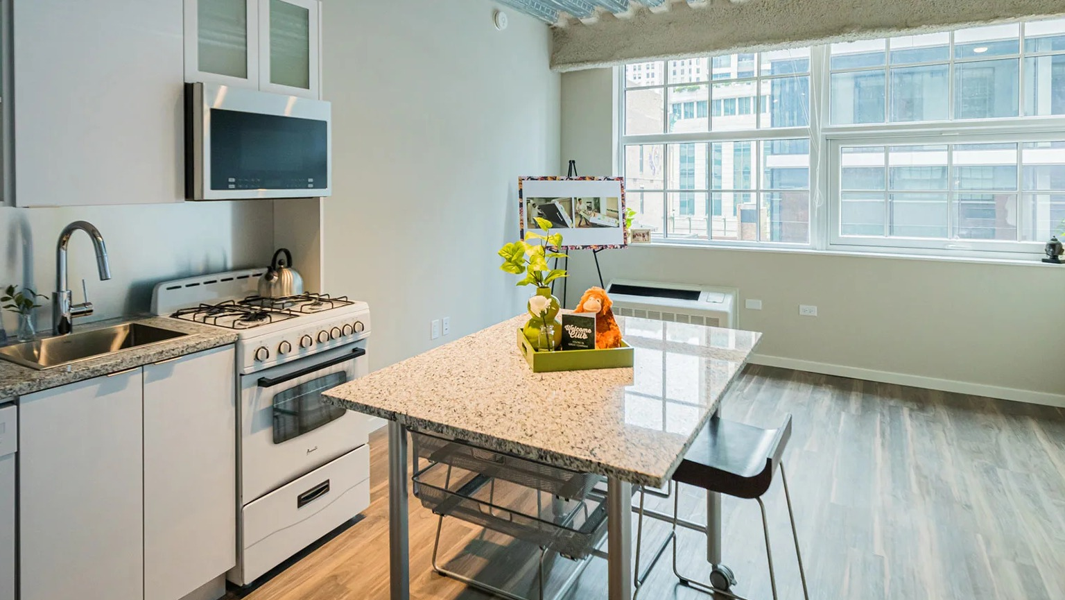 Compact kitchenette with white cabinets, a stove, microwave, and a dining table by a large window at MDA Apartments in Chicago