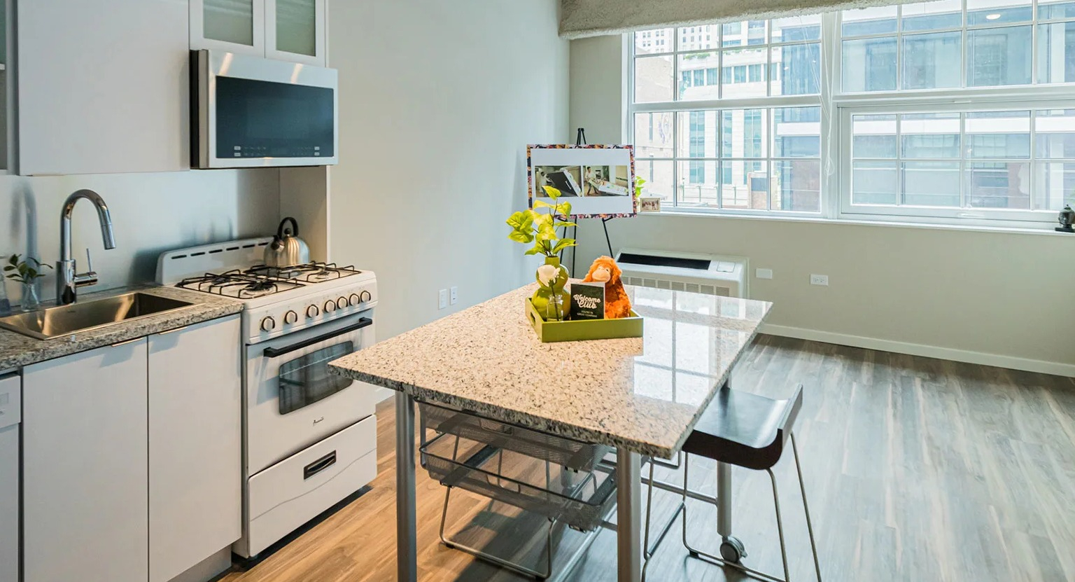 Compact kitchenette with white cabinets, a stove, microwave, and a dining table by a large window at MDA Apartments in Chicago