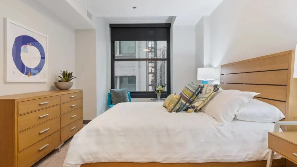 Bright bedroom featuring a wooden bed, dresser, and a large window with city views at Millennium on LaSalle in Chicago