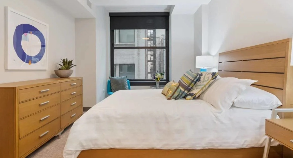 Bright bedroom featuring a wooden bed, dresser, and a large window with city views at Millennium on LaSalle in Chicago