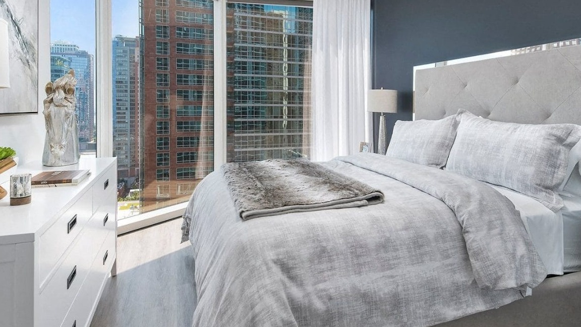 Cozy bedroom featuring a large upholstered bed with neutral-toned bedding and expansive windows showcasing city views at Moment Apartments in Chicago
