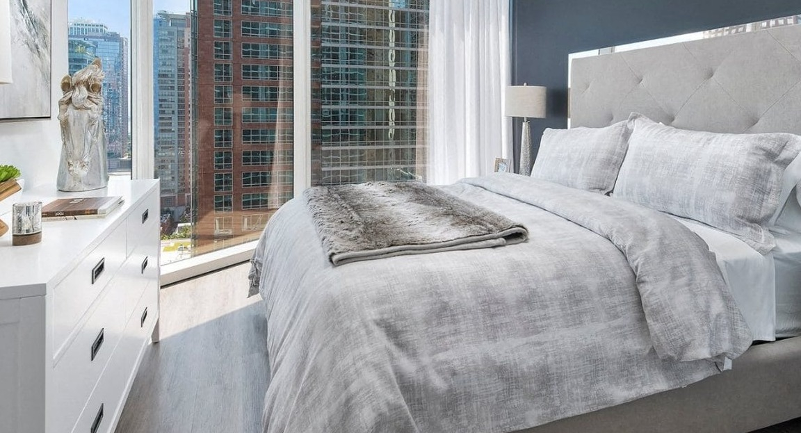 Cozy bedroom featuring a large upholstered bed with neutral-toned bedding and expansive windows showcasing city views at Moment Apartments in Chicago