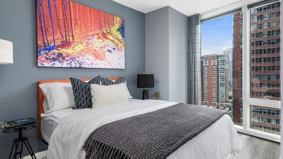 Modern bedroom with a vibrant abstract painting above the bed, featuring a large window with city views at Moment Apartments in Chicago