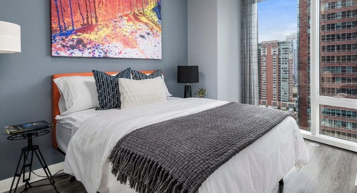 Modern bedroom with a vibrant abstract painting above the bed, featuring a large window with city views at Moment Apartments in Chicago