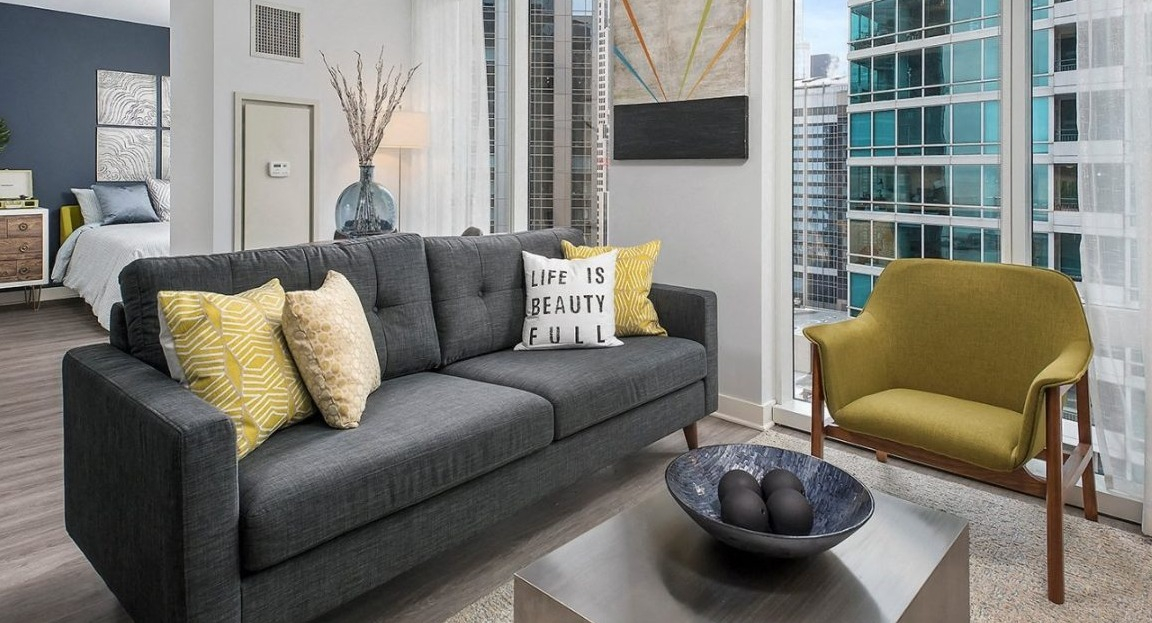Cozy living room with a dark gray sofa, yellow accent pillows, a modern coffee table, and large windows at Moment Apartments in Chicago