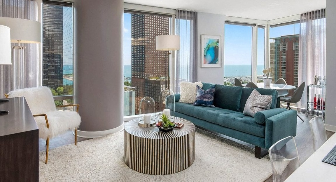 Spacious living room with a teal sofa, round coffee table, and expansive windows offering city and lake views at Moment Apartments in Chicago