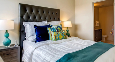 Cozy bedroom featuring a dark tufted headboard, vibrant blue and green pillows, and an open doorway to a bathroom at Mondial River West in Chicago
