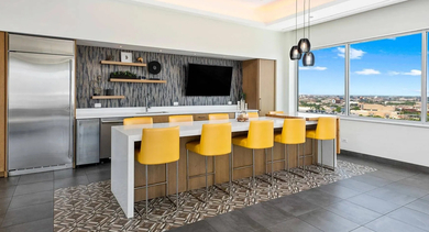 Spacious party room with a communal kitchen island, vibrant yellow stools, and large windows offering urban views at Mondial River West in Chicago