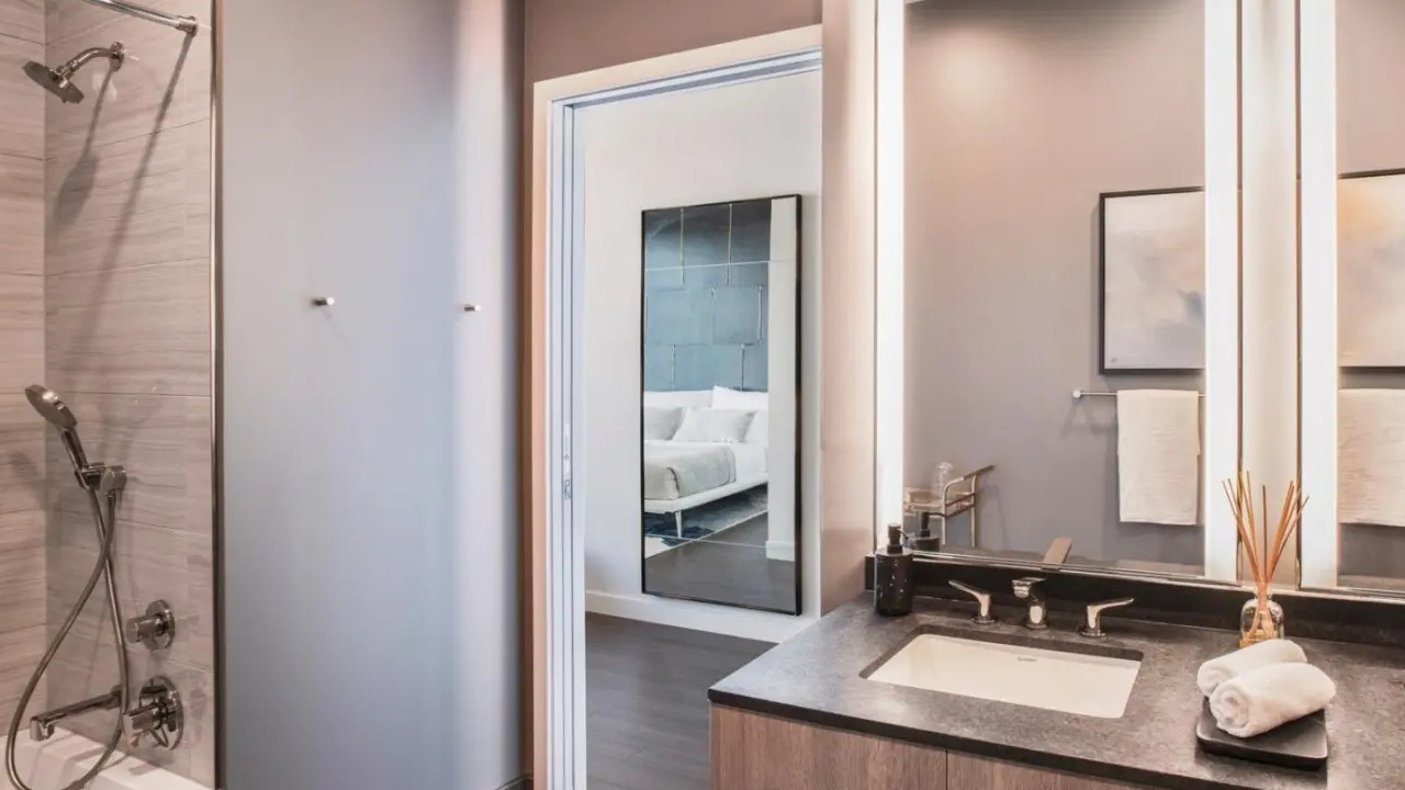 Luxurious apartment bathroom at NEMA Chicago, featuring a double vanity, illuminated mirrors, a modern shower, and a glimpse into the bedroom