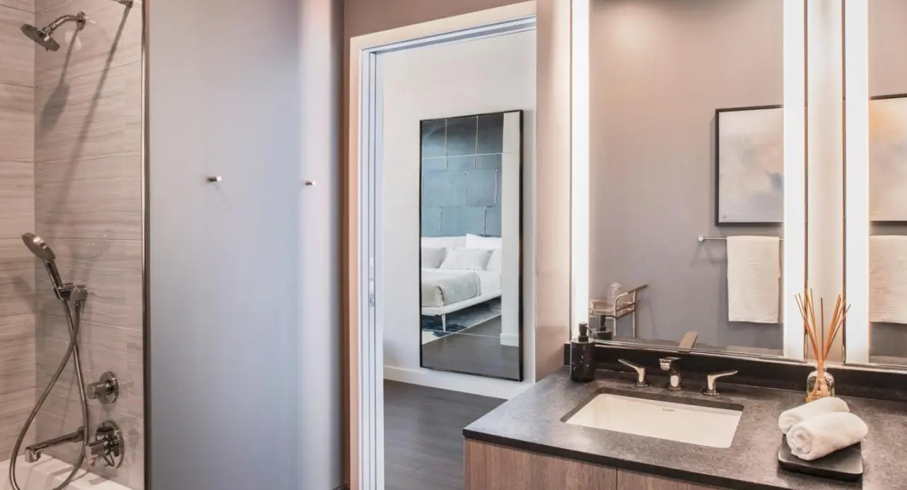 Luxurious apartment bathroom at NEMA Chicago, featuring a double vanity, illuminated mirrors, a modern shower, and a glimpse into the bedroom