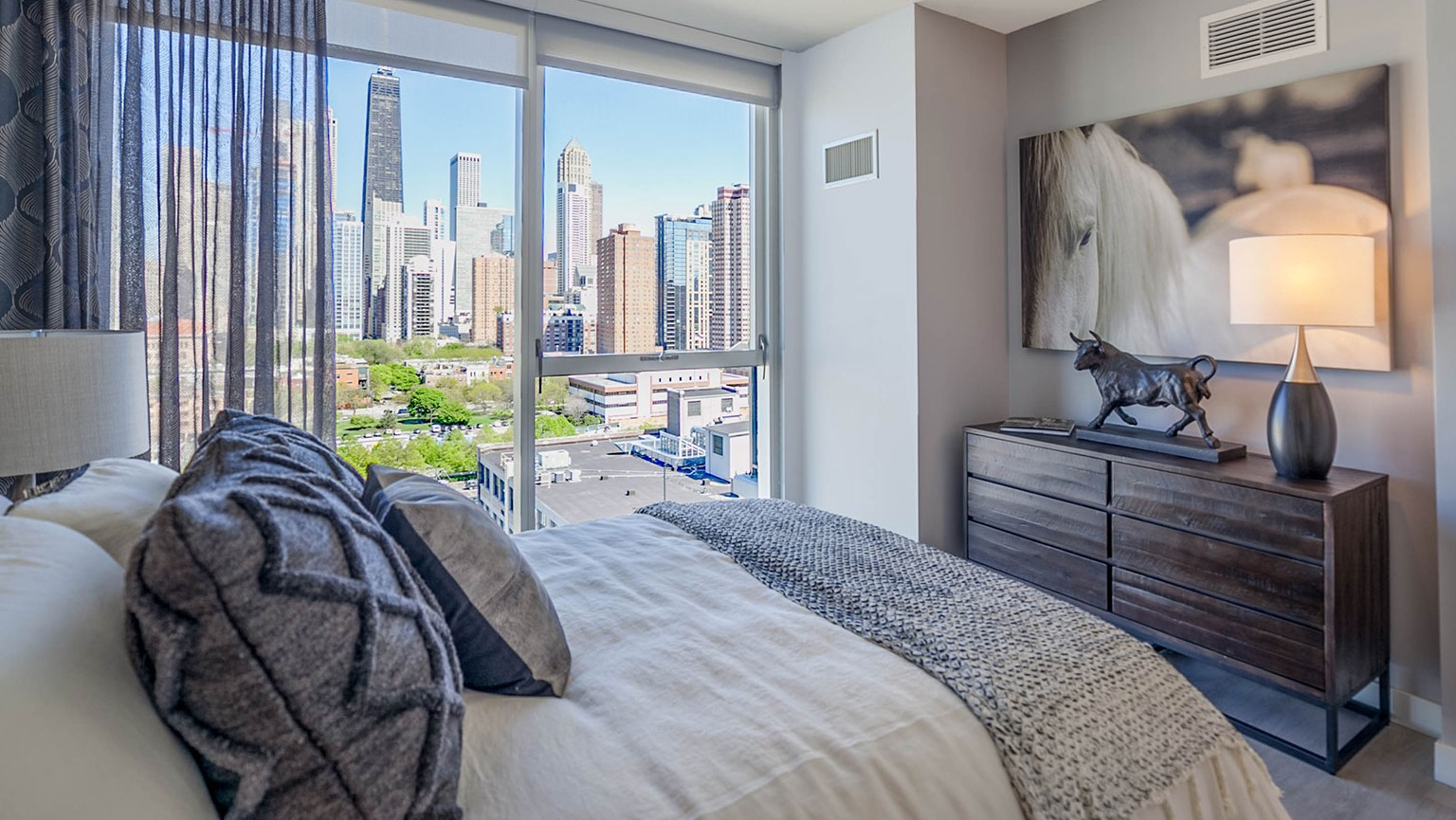 Elegant bedroom at Niche 905 in Chicago, featuring a comfortable bed, stylish dresser, and expansive city views through large windows