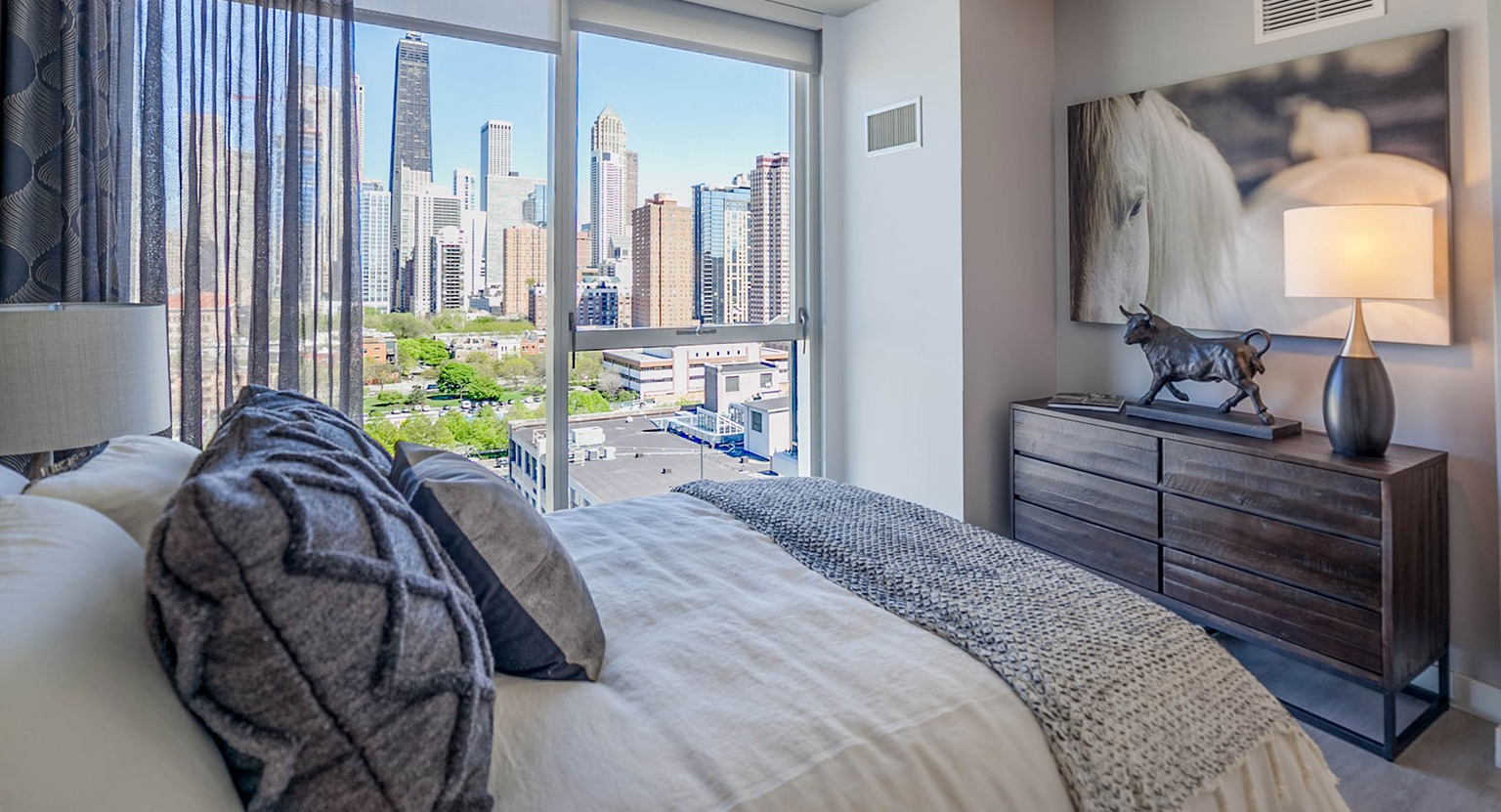 Elegant bedroom at Niche 905 in Chicago, featuring a comfortable bed, stylish dresser, and expansive city views through large windows