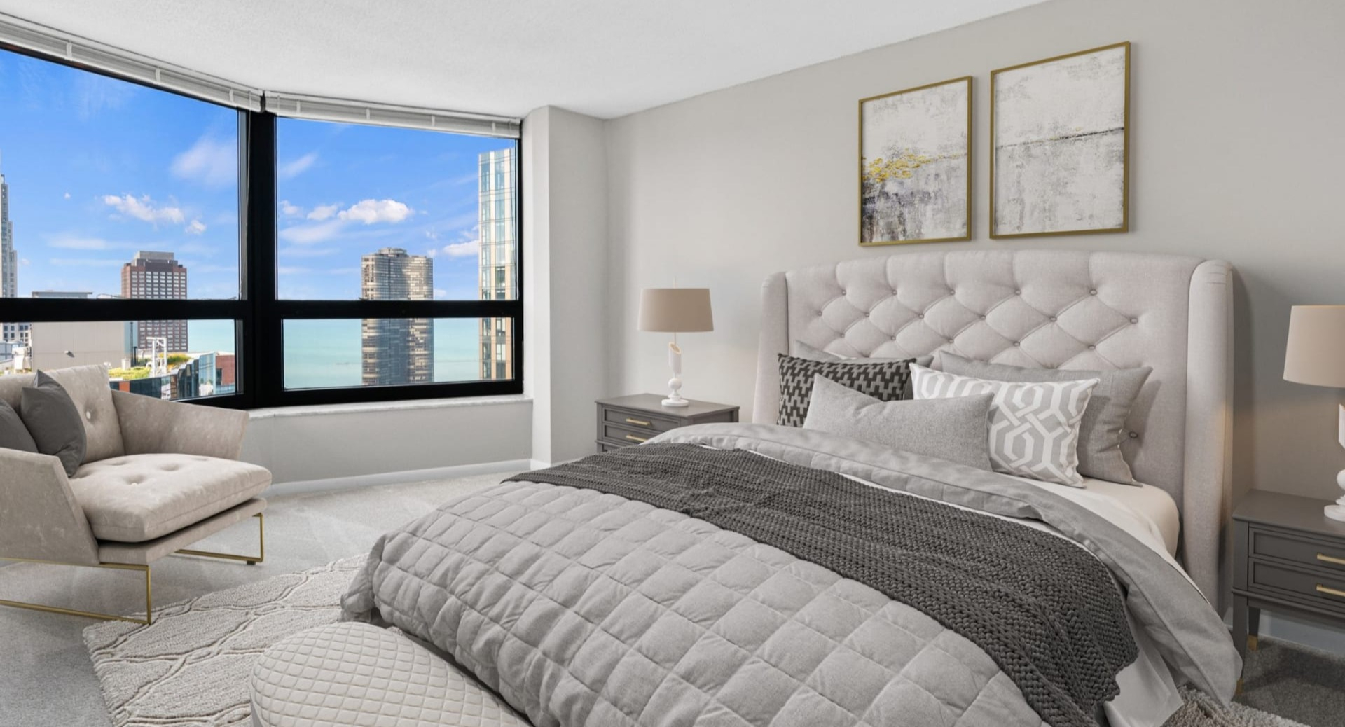 Sunlit bedroom corner with an upholstered bed, wall-mounted sconces, modern artwork, and large windows showcasing downtown city views at North Harbor Tower in Chicago