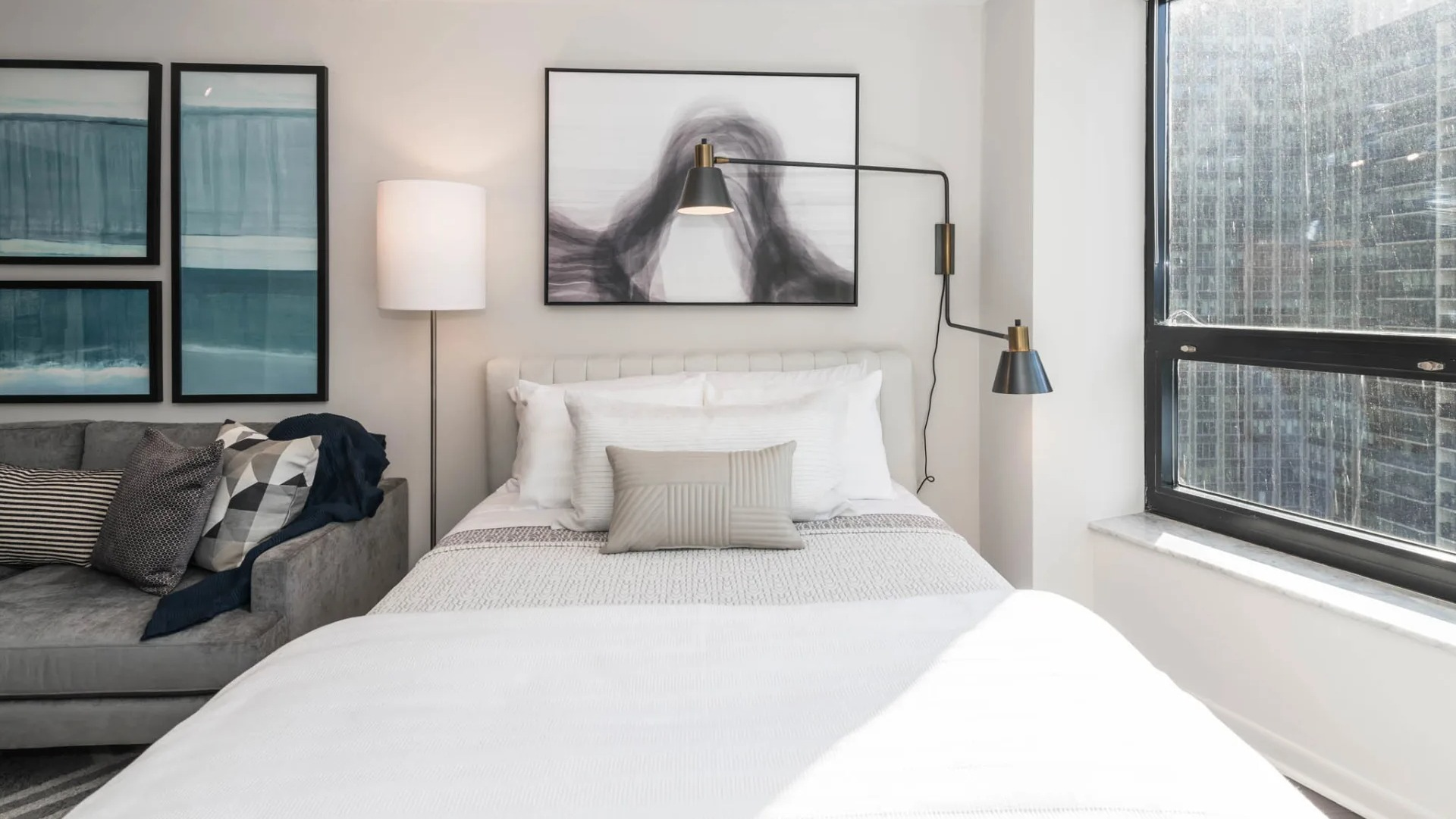 Cozy bedroom area at North Harbor Tower in Chicago with a plush bed, sofa seating, wall art, modern swing-arm lights, and a large window showcasing downtown views