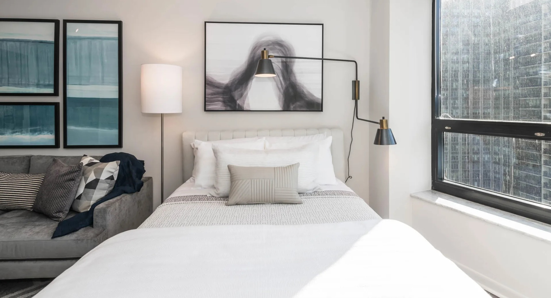 Cozy bedroom area at North Harbor Tower in Chicago with a plush bed, sofa seating, wall art, modern swing-arm lights, and a large window showcasing downtown views