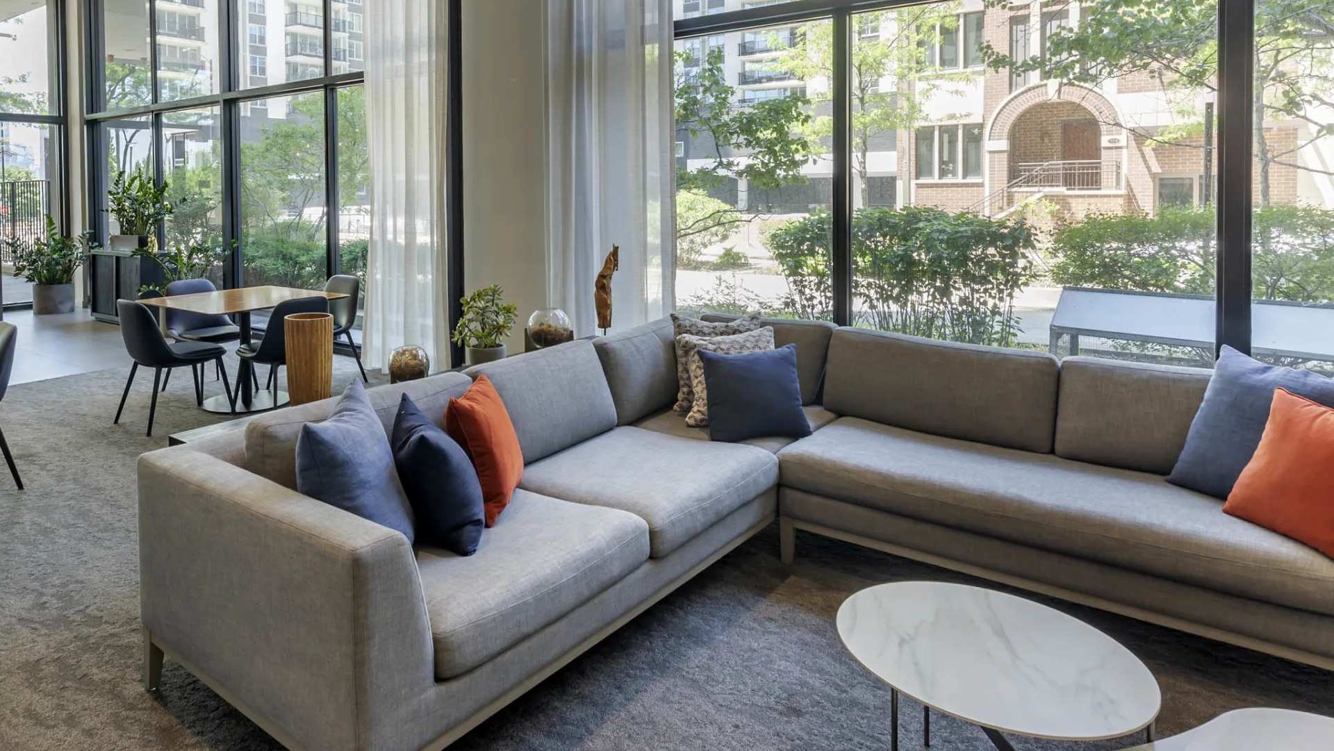 Spacious resident lounge at North Harbor Tower in Chicago with floor-to-ceiling windows, L-shaped sofa, accent pillows, and dining areas overlooking a tree-lined street